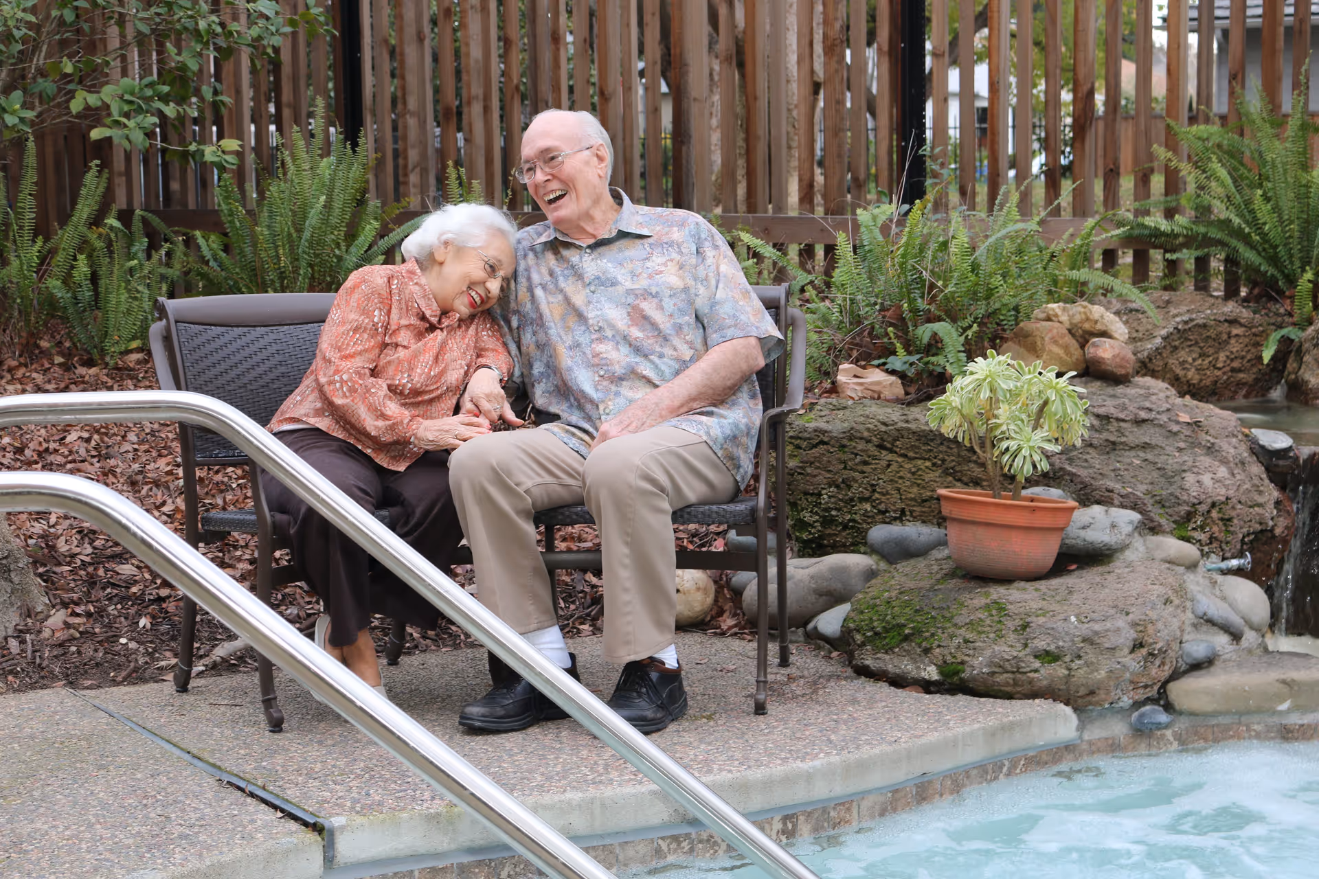 An elderly couple sitting on a bench beside a pool, smiling and holding hands. They are outdoors with a wooden fence, plants, and rocks in the background.