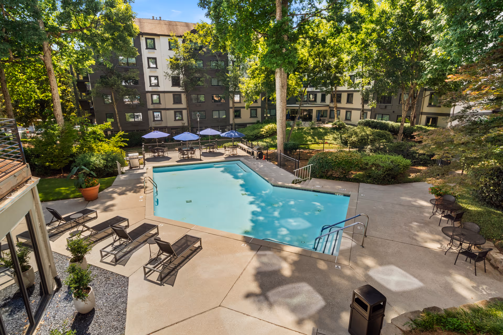 Outdoor swimming pool area surrounded by lounge chairs, tables with umbrellas, and lush green trees. In the background, there is a multi-story residential building with numerous windows.