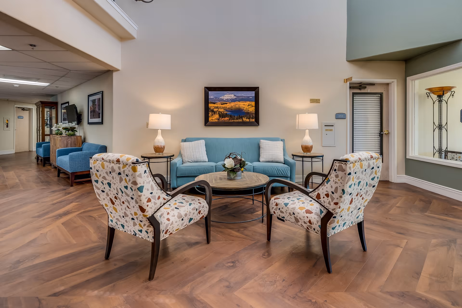 A cozy senior living facility common area with a light blue sofa, two patterned armchairs, a round wooden coffee table with a small flower arrangement, and two side tables with lamps. The walls are light-colored, and there is a framed landscape painting above the sofa. The floor is wood with a herringbone pattern. In the background, there are additional blue armchairs, a TV mounted on the wall, and a door labeled 'Office'.