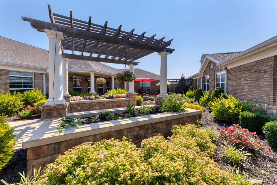 Outdoor garden area at Addington Place of Shoal Creek featuring a pergola with white columns, raised flower beds with various plants and flowers, and a patio with seating and a red umbrella under a clear blue sky.