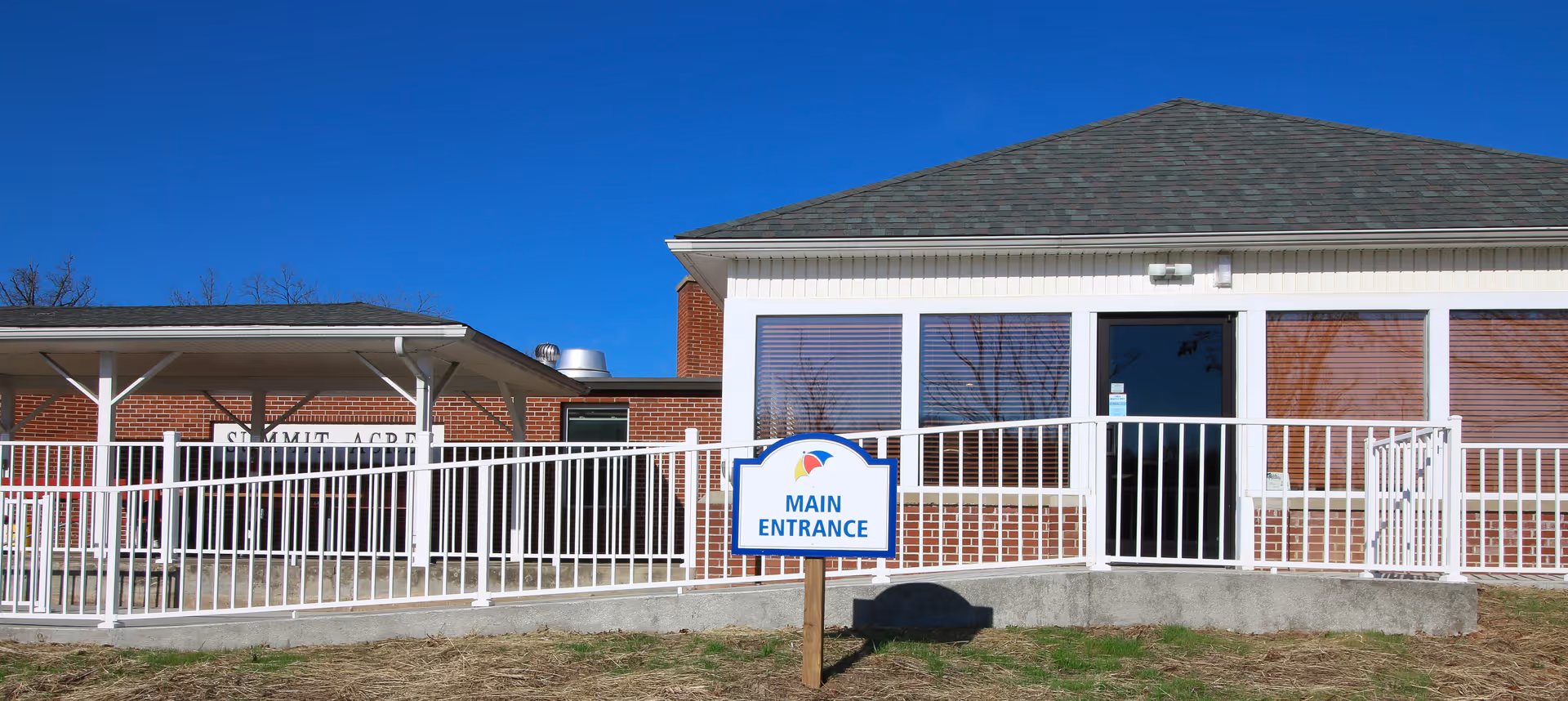 Front exterior of a single-story brick building with a ramp and a sign reading 'Main Entrance' under a clear blue sky.