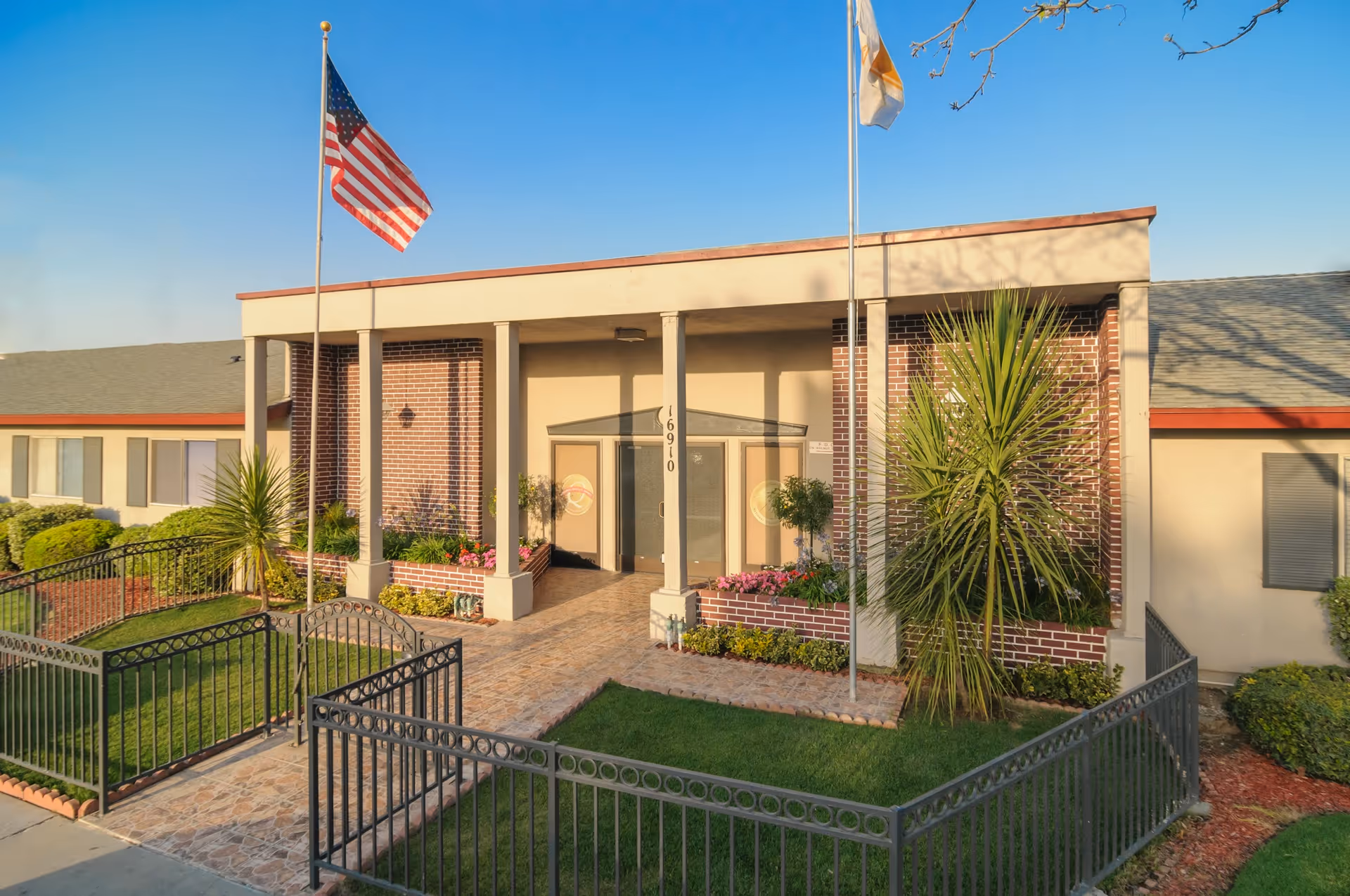 Front exterior view of Bel Tooren Villa Convalescent Hospital building with two flagpoles displaying the American flag and another flag, a gated entrance, well-maintained landscaping with green grass, bushes, and plants, and a clear blue sky.