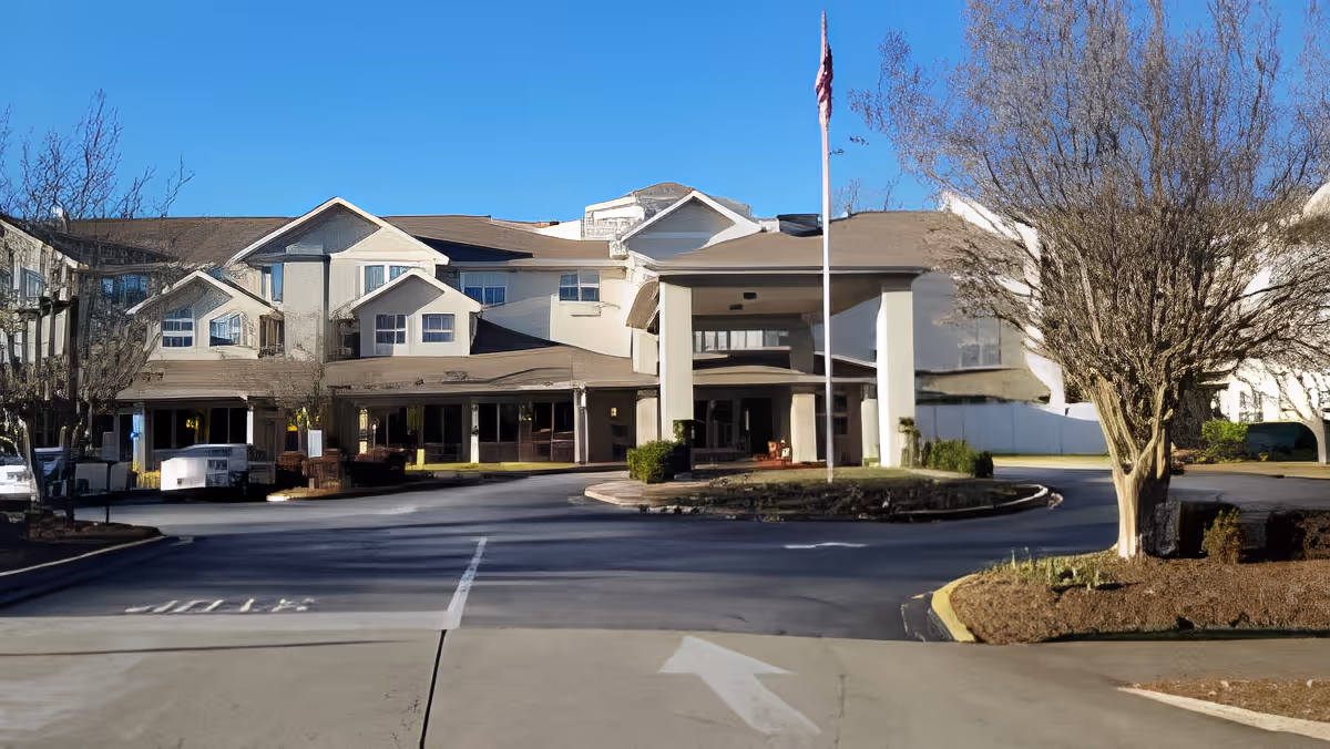 Front exterior view of a multi-story senior living facility building with a covered entrance, an American flag on a flagpole, leafless trees, and a driveway with directional arrows.
