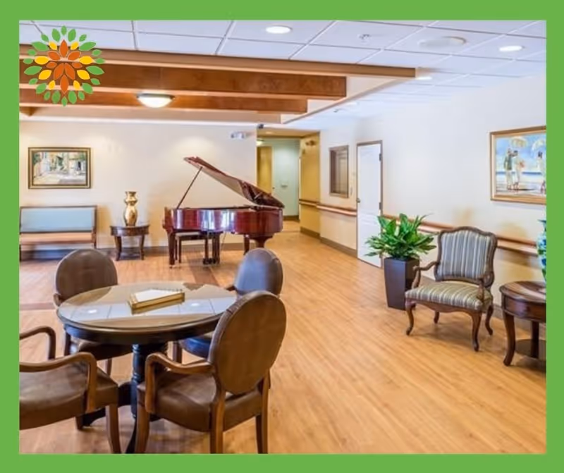 A spacious room with wooden flooring featuring a round table with four chairs in the foreground, a grand piano against the back wall, a bench, side tables with decorative items, framed paintings on the walls, and a potted plant near a striped armchair.