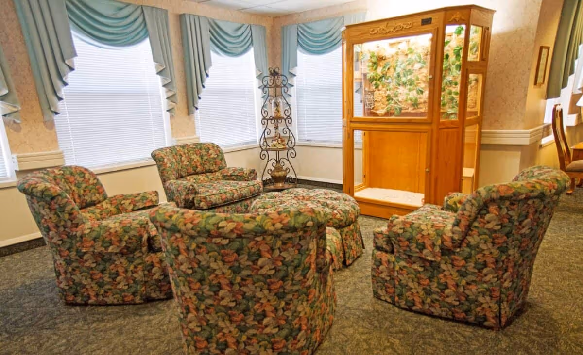 A cozy sitting area with four floral-patterned armchairs arranged around a matching ottoman. The room has three windows with teal valances and white blinds. There is a wooden display case with plants inside and a decorative metal stand with small ornaments in the corner.