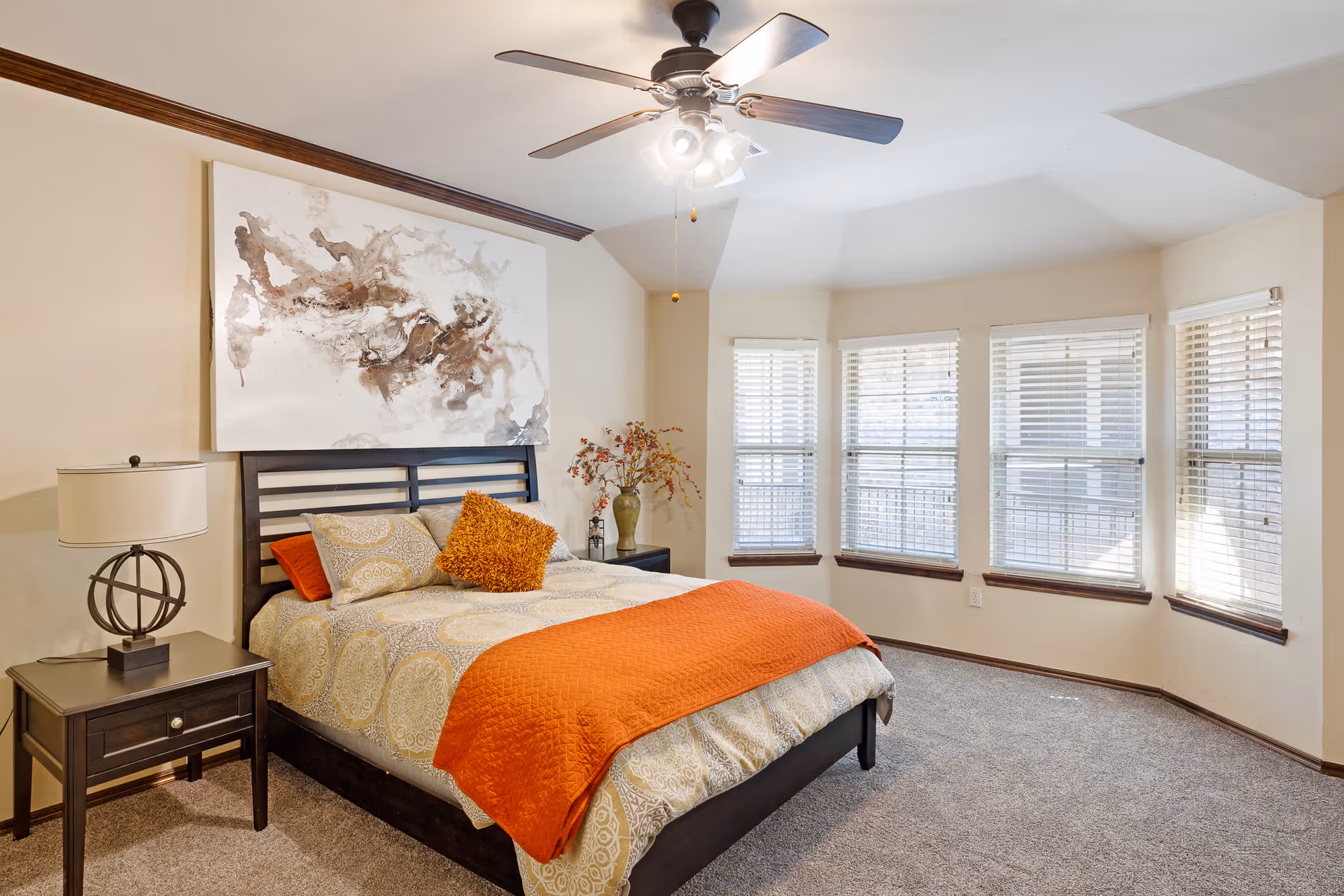 A cozy bedroom with a large bed featuring patterned bedding and an orange throw blanket. There is a nightstand with a modern lamp on the left side of the bed. Above the bed hangs an abstract painting. The room has a ceiling fan with lights and a set of five windows with blinds letting in natural light. The floor is carpeted and the walls are painted a light beige color.