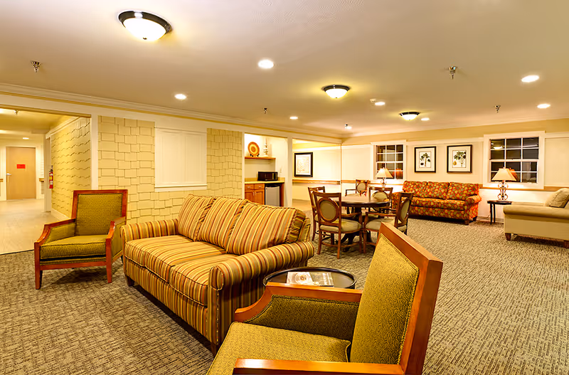 Well-lit communal lounge with a striped sofa, armchairs, a round dining table, and additional seating and lamps.