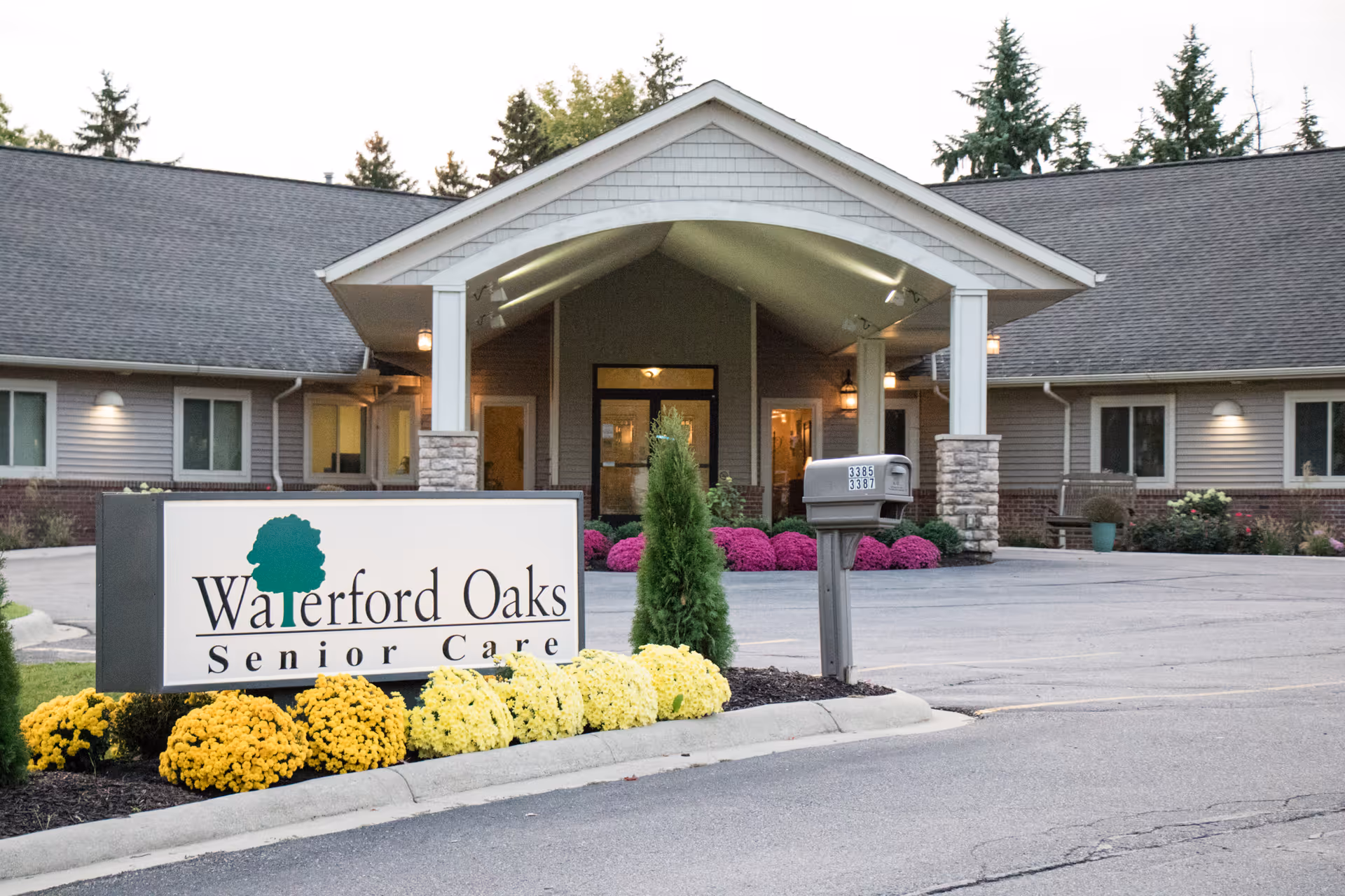 Exterior front view of Waterford Oaks Senior Care facility with a covered entrance, a sign displaying the facility name, a mailbox, and landscaped flowers in front.