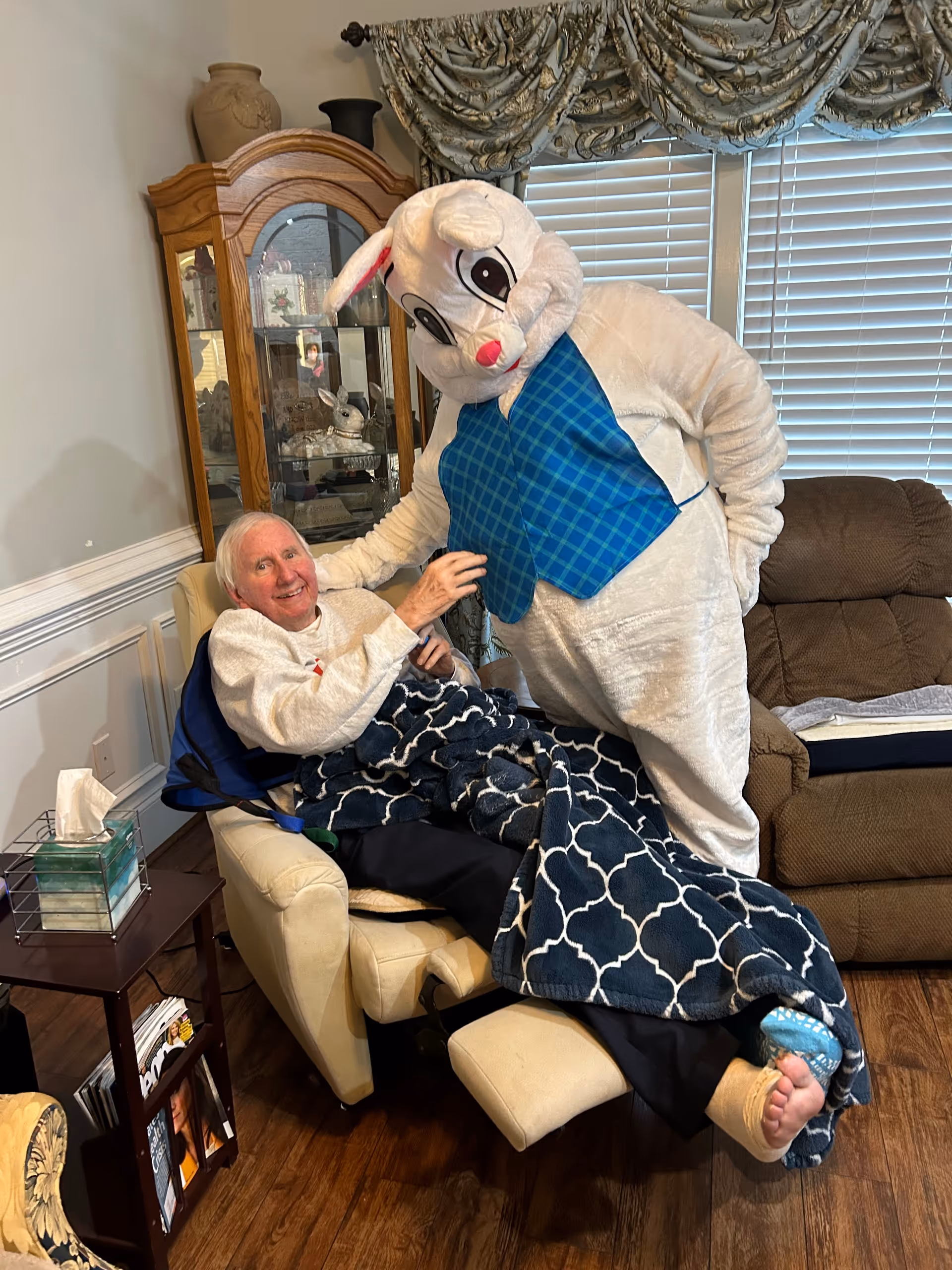A smiling older adult reclines in a chair covered by a patterned blanket as a person in a large white bunny costume leans in beside them in a living room.