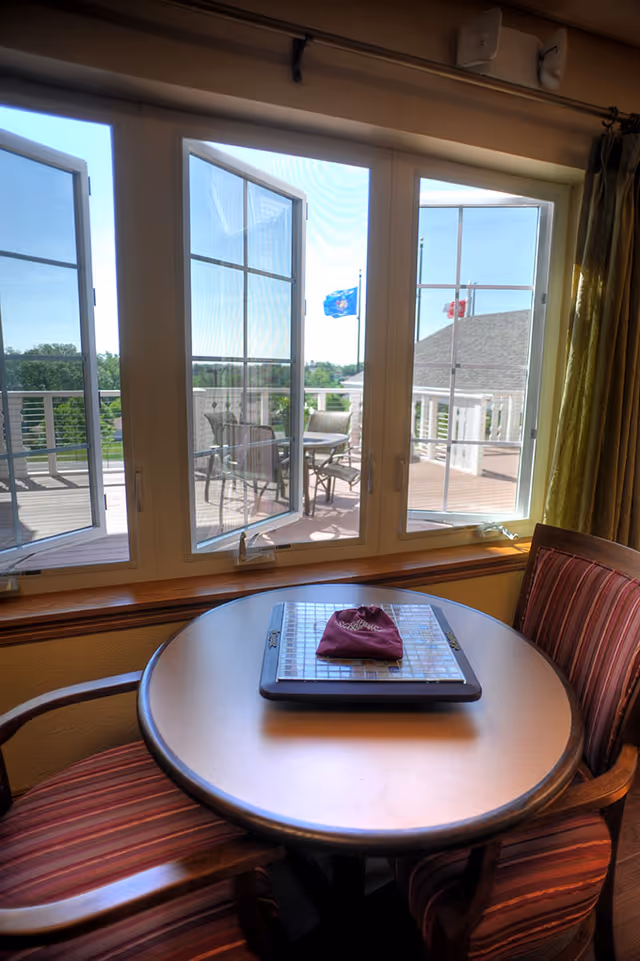 Round table with two striped chairs sits by large windows overlooking a sunlit outdoor deck with patio furniture and flags.