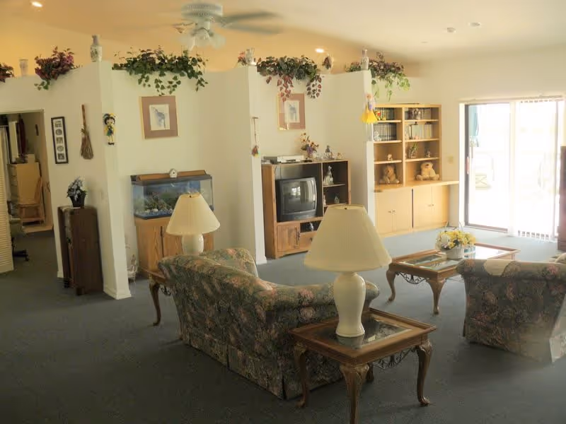 A cozy living room area with floral patterned sofas, two table lamps on wooden side tables, a coffee table with a flower arrangement, a TV on a wooden stand, a fish tank, a bookshelf with teddy bears, and sliding glass doors letting in natural light. The walls are decorated with framed pictures and plants on top of a partial wall divider.