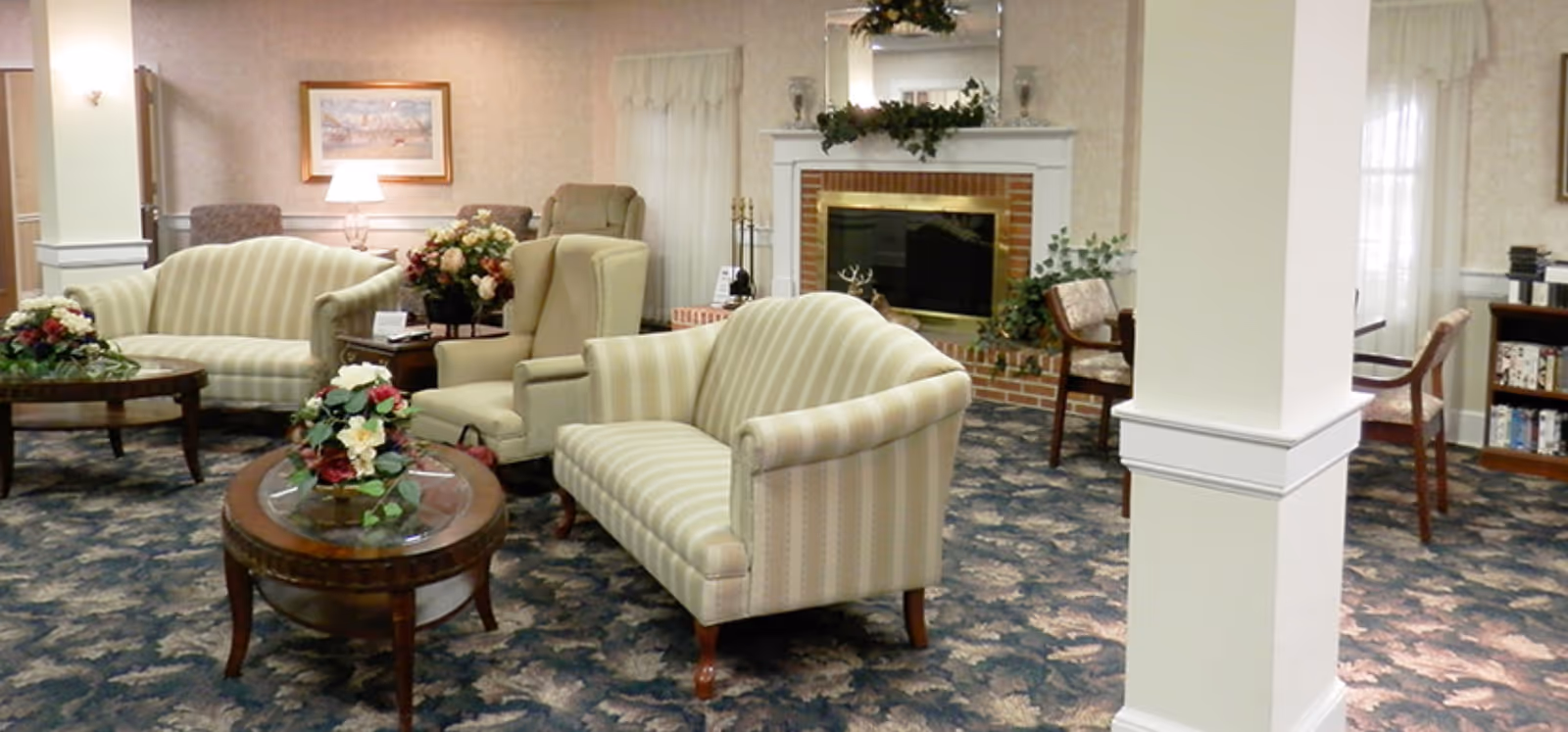 A cozy living room area in Colonial Gardens featuring striped upholstered sofas and armchairs arranged around wooden coffee tables with floral centerpieces. The room has a patterned carpet, a brick fireplace with a white mantel decorated with greenery and candles, and framed artwork on the walls. There are also windows with sheer curtains and a bookshelf in the background.