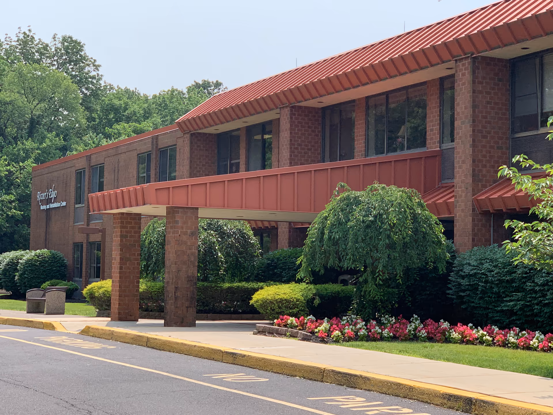 Front entrance of River's Edge Rehabilitation and Healthcare Center, a two-story brick building with a covered drive-through, shrubs and flowerbeds.