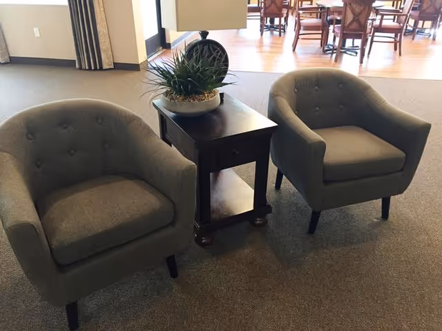 Two gray upholstered armchairs with button tufting are positioned on either side of a dark wooden side table with a drawer and lower shelf. A potted plant with green spiky leaves sits on top of the table. In the background, there is a dining area with wooden chairs and tables on a hardwood floor.