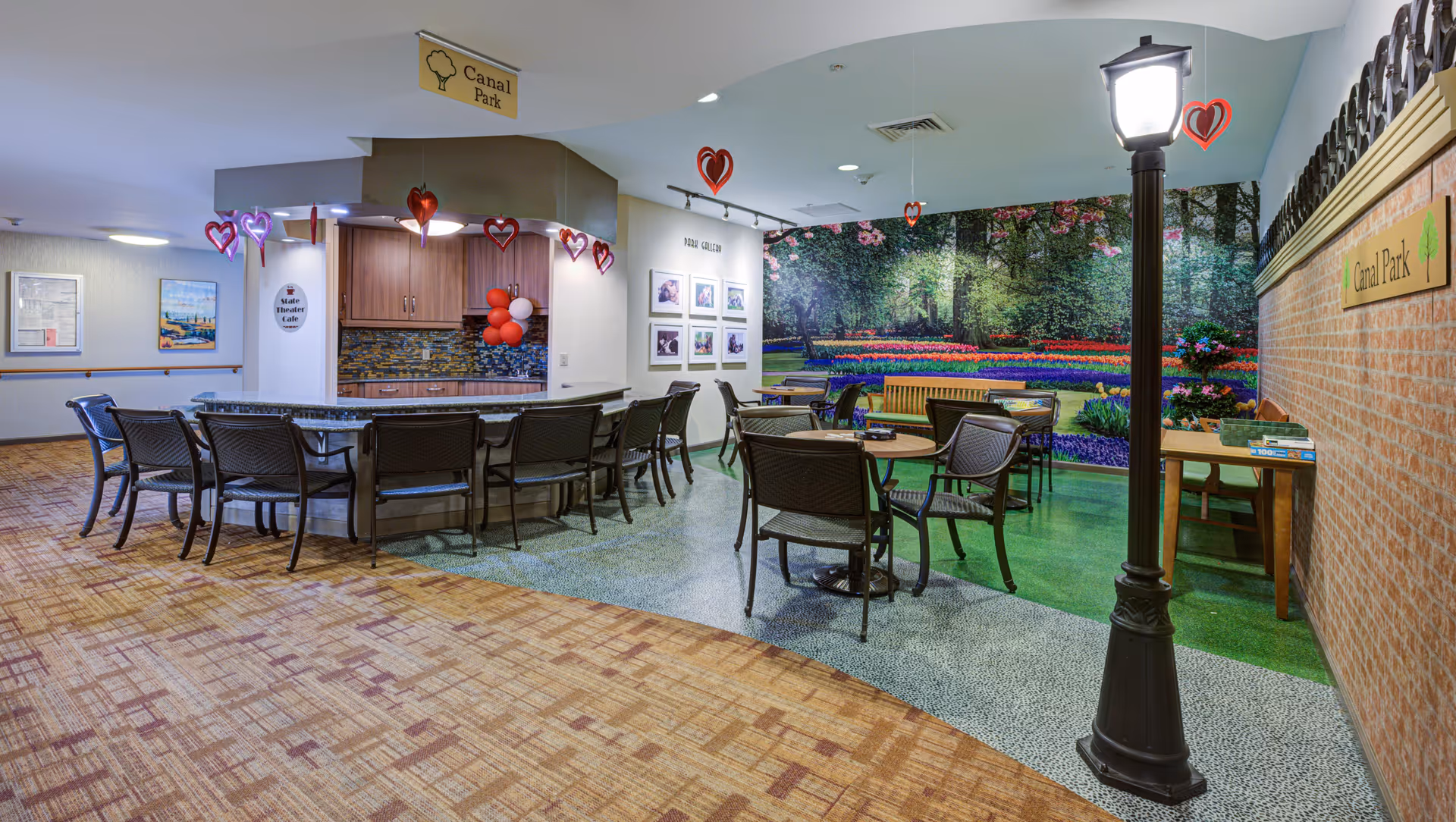 Interior common area of Arden Courts - ProMedica Memory Care Community with a seating area featuring tables and chairs, a counter with bar stools, a decorative street lamp, and a large mural of a colorful garden on the wall. Heart-shaped decorations hang from the ceiling.