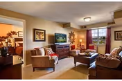 A cozy living room in a retirement community featuring a sofa, armchairs, a wooden coffee table, and a TV on a wooden stand. The room has beige walls, carpeted floor, and a window with red curtains allowing natural light to enter. Decorative items and framed pictures adorn the walls and furniture.