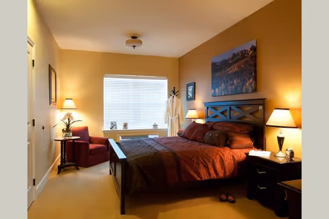 A warmly lit bedroom with a large bed featuring dark wood frame and red bedding. There are two bedside tables with lamps, a red armchair next to a small round table with a plant, and a window with blinds letting in natural light. A robe hangs on a coat rack near the window, and a landscape painting is mounted above the bed's headboard.