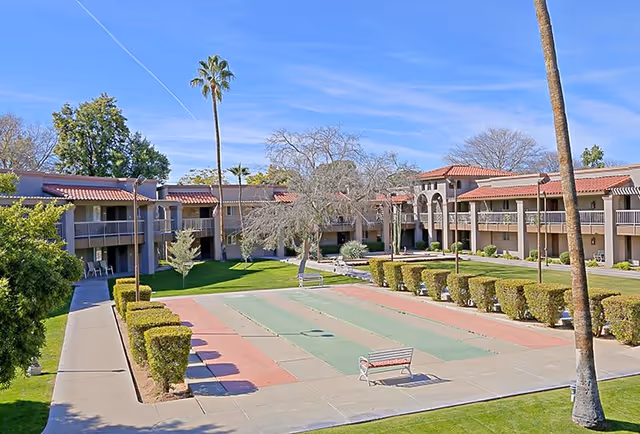 Outdoor courtyard area of a senior living facility with a shuffleboard court in the center, surrounded by trimmed bushes and palm trees. The two-story building with balconies and red-tiled roofs encloses the courtyard under a clear blue sky.