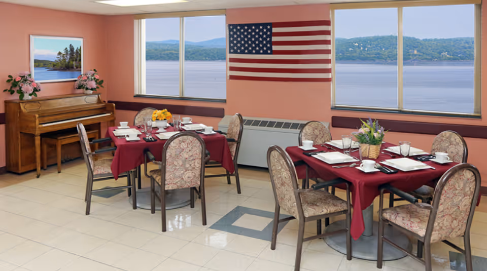 Dining room with two tables covered in burgundy tablecloths, each set with white plates, cups, and silverware. Six floral upholstered chairs surround the tables. An American flag hangs on the wall between two large windows overlooking a body of water and distant hills. A wooden piano with flower arrangements on top is positioned against the left wall beneath a framed picture of a lake scene.