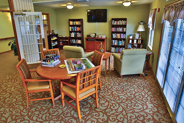 A comfortable common area with bookshelves, a table, and seating in a senior living facility.
