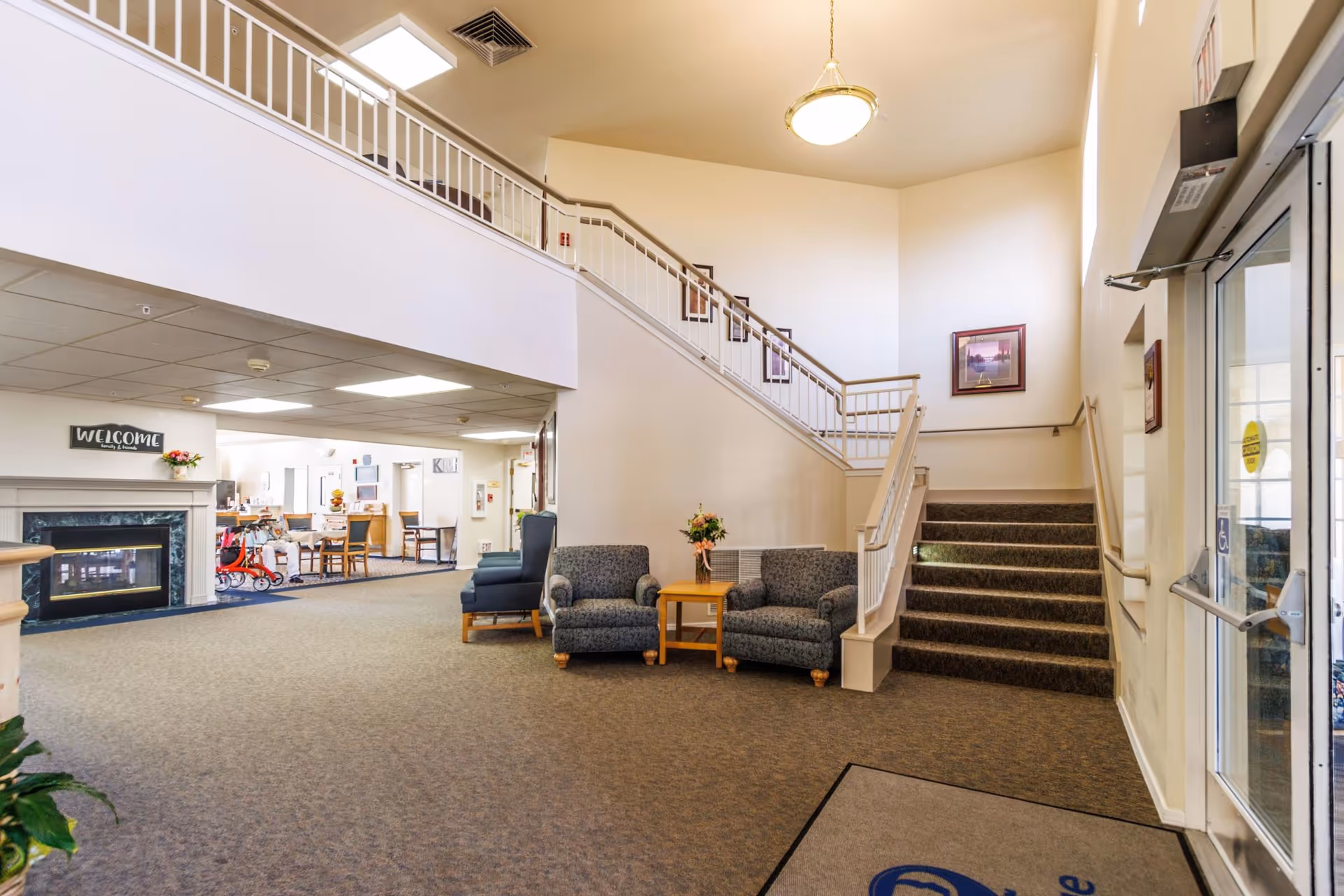 Interior view of a senior living facility lobby with a staircase leading to an upper floor. The area includes two patterned armchairs with a small wooden table and flowers between them, a blue armchair, and a fireplace with a 'WELCOME family & friends' sign above it. In the background, there is a dining area with tables and chairs. The space is well-lit with ceiling lights and a hanging light fixture.