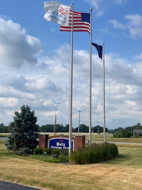 Three flagpoles with flags flying in the wind, including an American flag, standing behind a sign that reads 'Betz Nursing Home' surrounded by grass and a small tree under a partly cloudy sky.