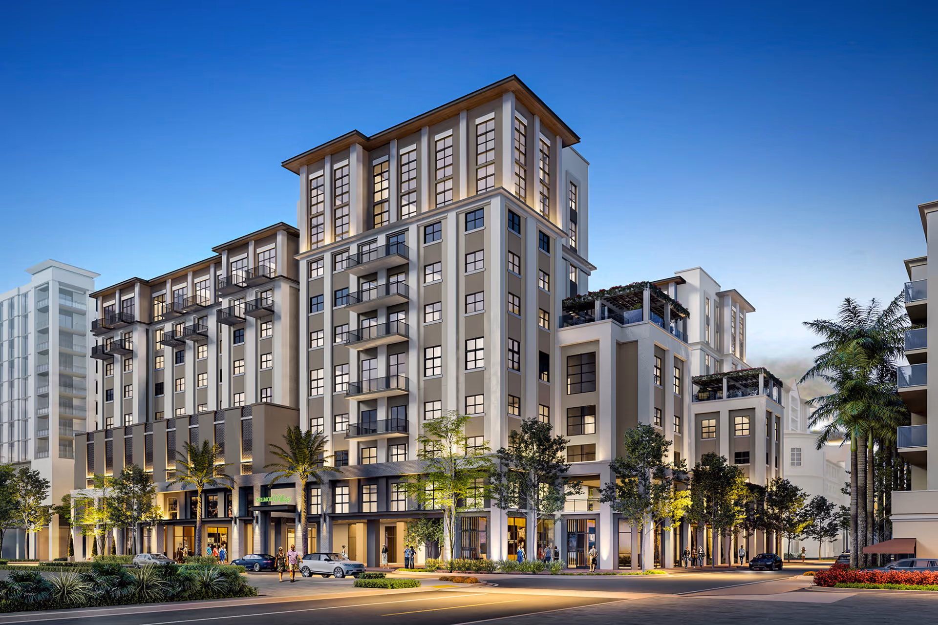 Exterior view of a modern multi-story residential building at dusk with illuminated windows and balconies, surrounded by palm trees and landscaped greenery. People and cars are visible near the entrance and along the street.