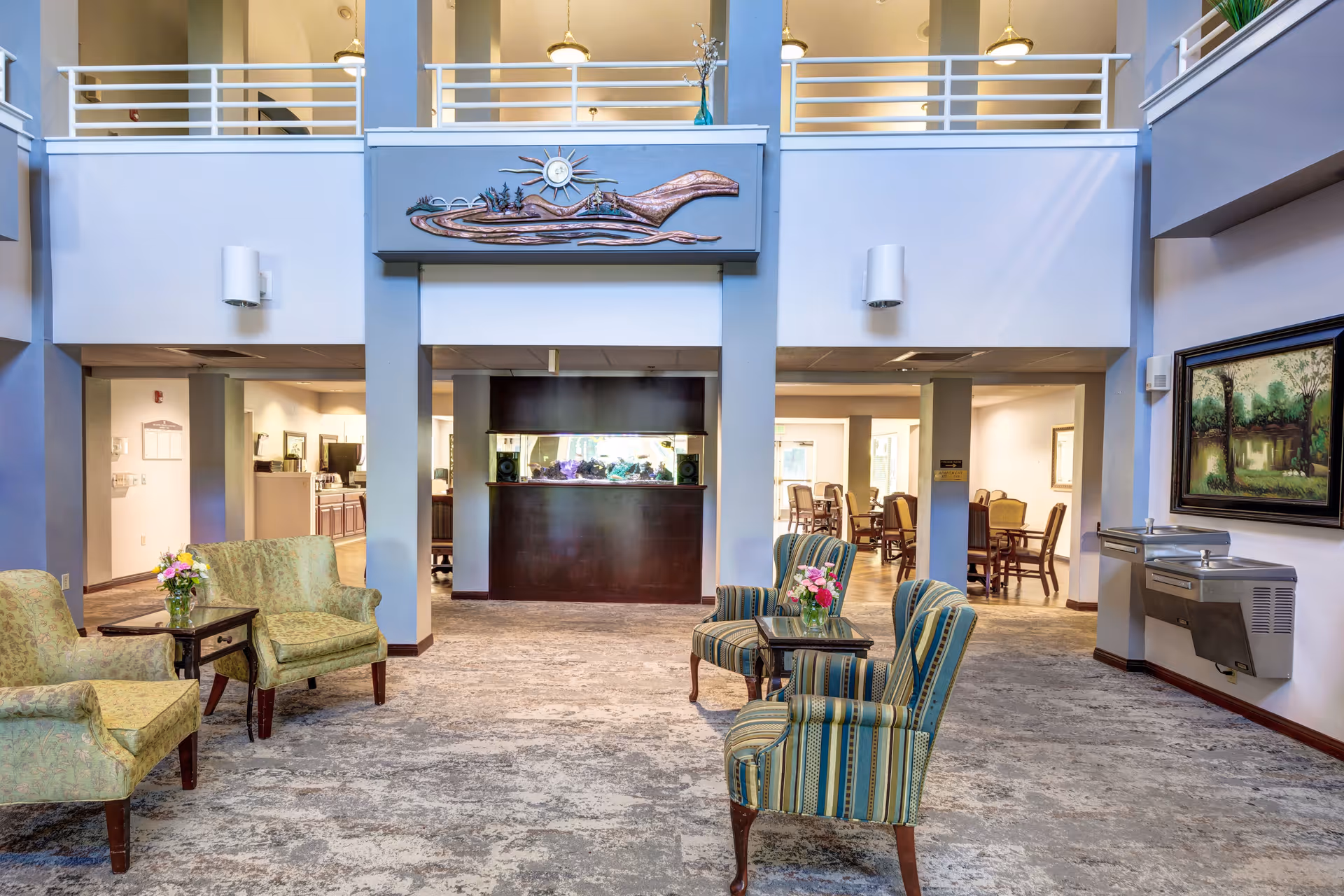 A spacious and well-lit common area in an assisted living facility featuring comfortable armchairs with floral and striped upholstery arranged around small tables with flower vases. The room has a high ceiling with a balcony railing above, a decorative wall art piece, and a water fountain on the right wall. In the background, there is a dining area with tables and chairs.