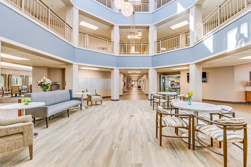 Bright and spacious common area in an assisted living facility with light wood flooring, modern seating including a gray sofa and patterned chairs, round tables with small flower vases, and a high ceiling with hanging light fixtures. The area opens into a long hallway and adjacent rooms with additional seating and dining spaces.