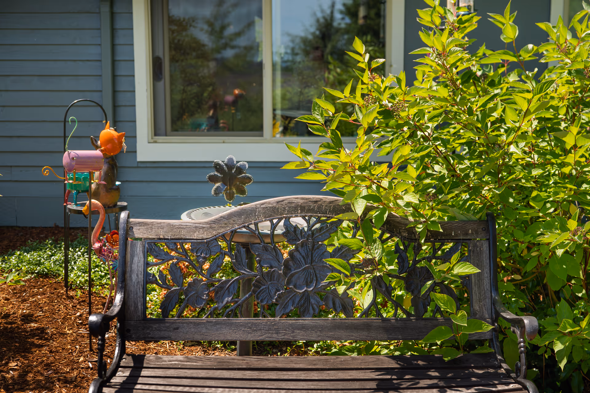 A decorative wooden bench with floral metalwork is placed outdoors in a garden area with green leafy bushes and a blue building wall with a window in the background. There is also a colorful metal garden ornament shaped like a cat and a flamingo near the bench.