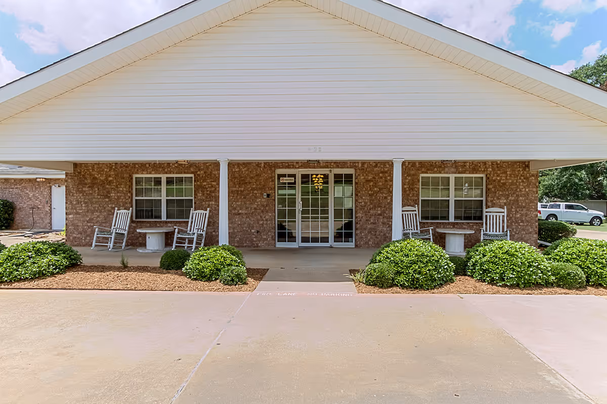 Front exterior view of a senior living facility with a covered entrance supported by white columns. There are two sets of white rocking chairs and small round tables on either side of the glass double doors. The building has a brick facade with two windows visible, surrounded by neatly trimmed bushes and a concrete driveway in front.