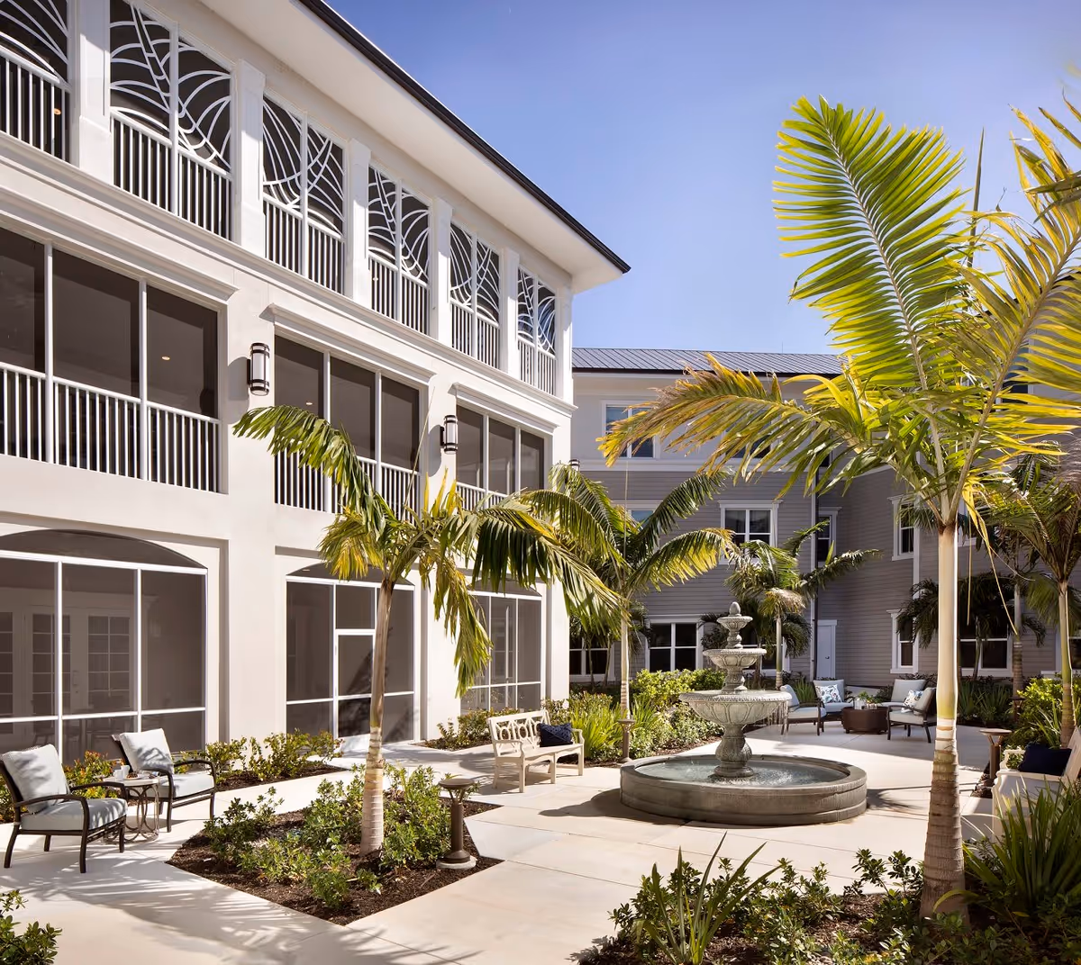Outdoor courtyard area at The Preserve in Fort Myers featuring a central stone fountain surrounded by palm trees, benches, and patio chairs. The building has large screened windows and decorative upper window panels under a clear blue sky.
