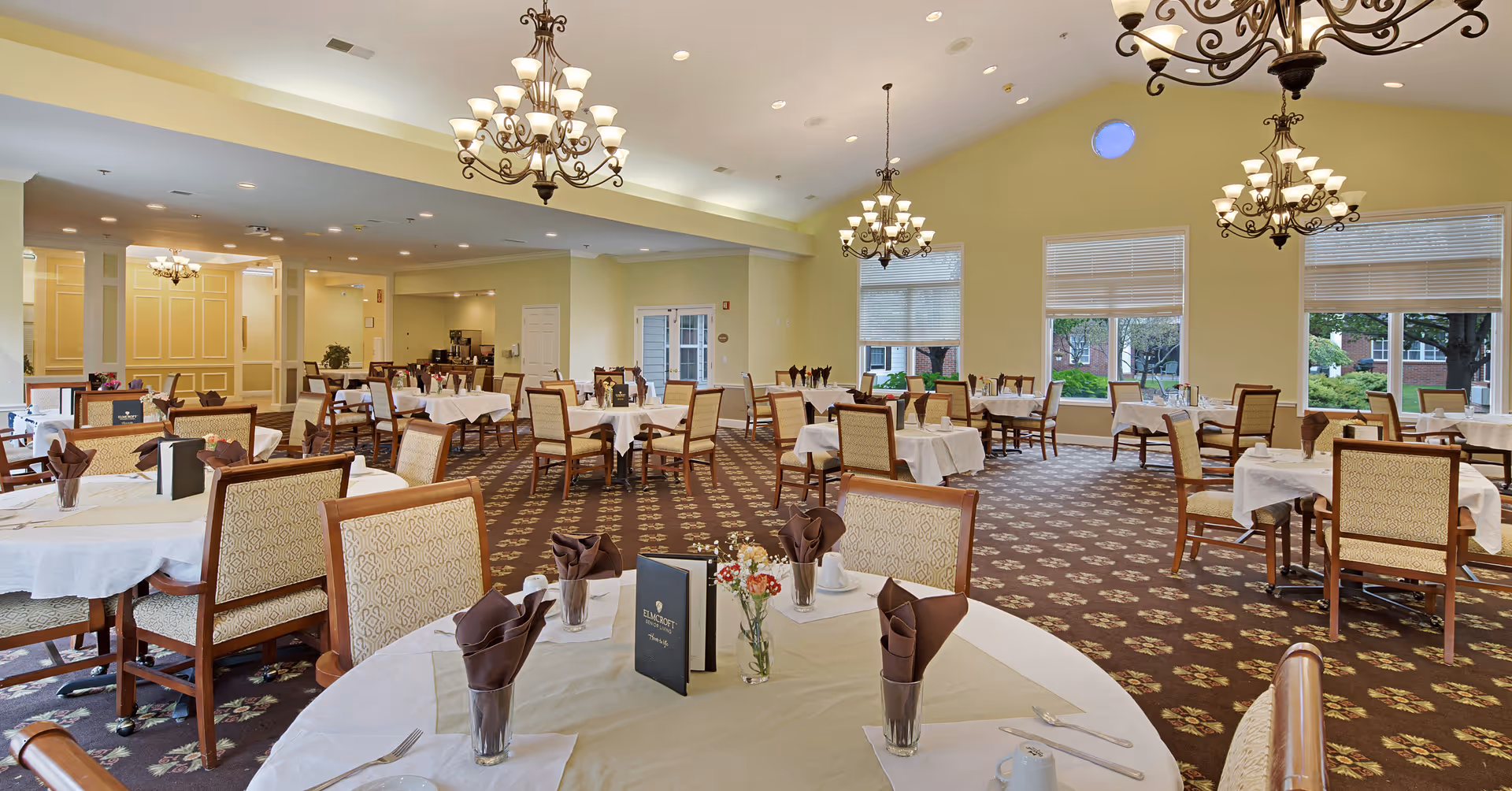 A spacious dining room with multiple round and square tables covered with white tablecloths, each set with brown napkins, glassware, and menus. The room features large windows letting in natural light, patterned carpet, and elegant chandeliers hanging from the ceiling.