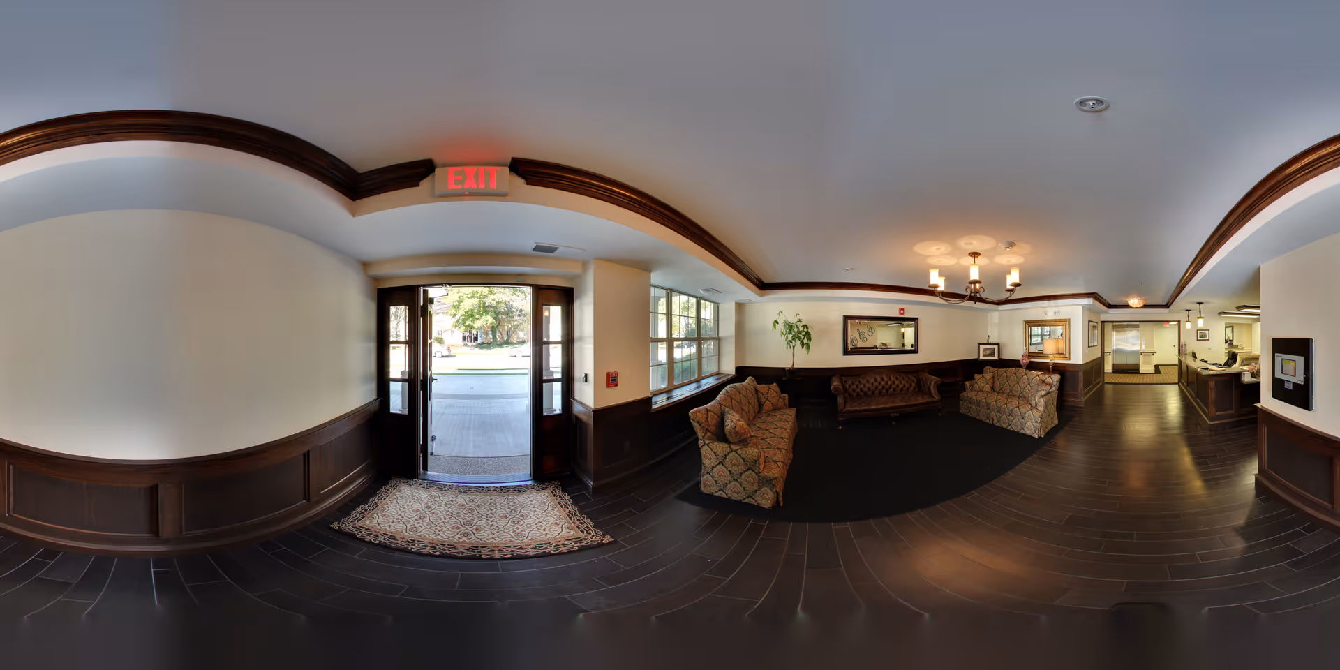 Interior view of a senior living facility lobby with dark wood paneling and flooring, a glass door exit leading outside, patterned upholstered sofas arranged around a dark rug, a large window letting in natural light, a chandelier hanging from the ceiling, and a reception desk in the background.
