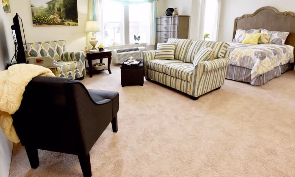 A cozy room with a bed, a striped loveseat, an armchair with a yellow blanket, a patterned armchair, a wooden dresser, a side table with a lamp, and a window with light blue curtains letting in natural light.