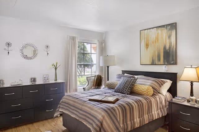 Sunlit bedroom with a striped bed, headboard, matching dresser and nightstand, a window with curtains, lamps, and wall art.