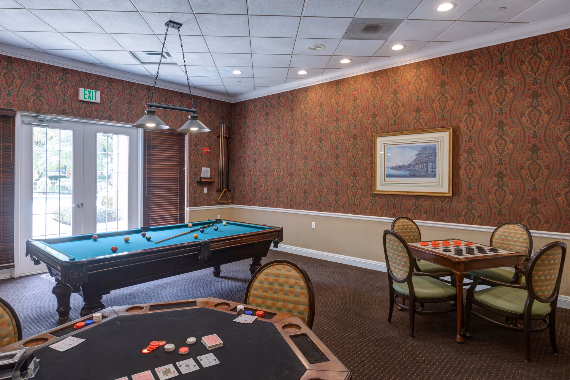 A game room with a pool table, a card table with poker chips and playing cards, and a checkers table with four chairs. The room has patterned wallpaper, a framed picture on the wall, and a glass door leading outside with an exit sign above it.