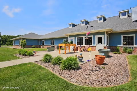 Exterior view of a single-story blue senior living building with a patio, landscaped beds, and potted plants under a blue sky.