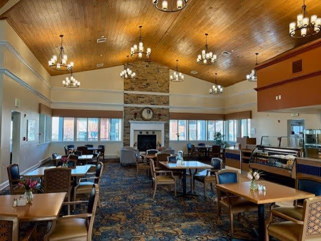 A spacious dining room with multiple tables and chairs arranged neatly. The room features a high wooden ceiling with several chandeliers, large windows letting in natural light, a stone fireplace with a clock above it, and a carpeted floor with a patterned design. There is a display case on the right side and some plants near the windows.