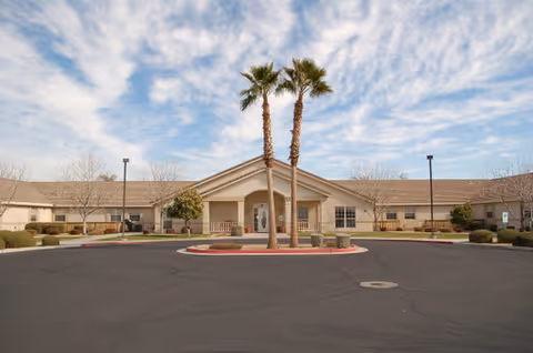 Front exterior view of a single-story building with a covered entrance, two tall palm trees in front, and a circular driveway. The sky is partly cloudy.