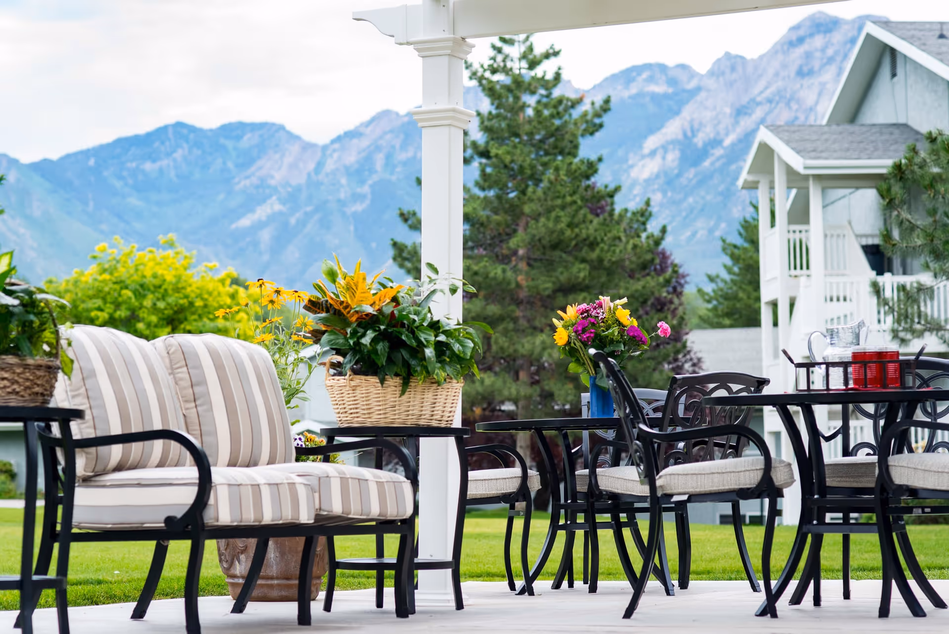 Outdoor patio area with cushioned striped loveseat, black metal chairs and tables, and colorful flower arrangements in baskets and vases, set against a backdrop of green trees and mountains under a cloudy sky.