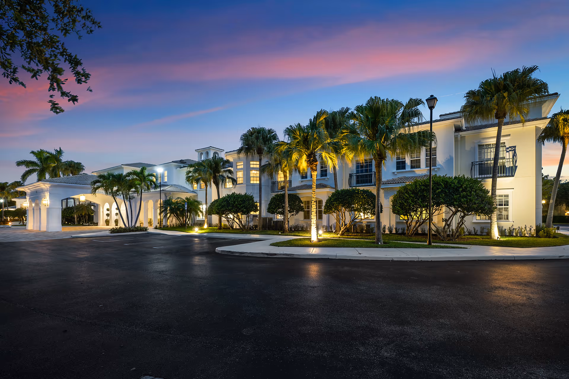 Exterior view of Tequesta Terrace senior living facility at dusk, showing a well-lit two-story building with palm trees and landscaped bushes along the sidewalk under a colorful sky.