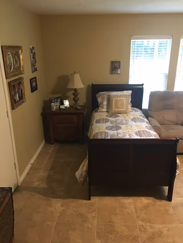 A bedroom with a single dark wooden bed featuring a patterned bedspread and two pillows. Next to the bed is a wooden nightstand with a lamp, framed photos, and small decorative items. On the wall above the nightstand are several framed pictures. A beige recliner chair is positioned near two windows with white blinds, allowing natural light into the room. The floor is tiled in a light brown color.