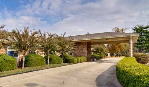 Driveway leading to a brick nursing center entrance with a covered porte-cochere and trimmed bushes.