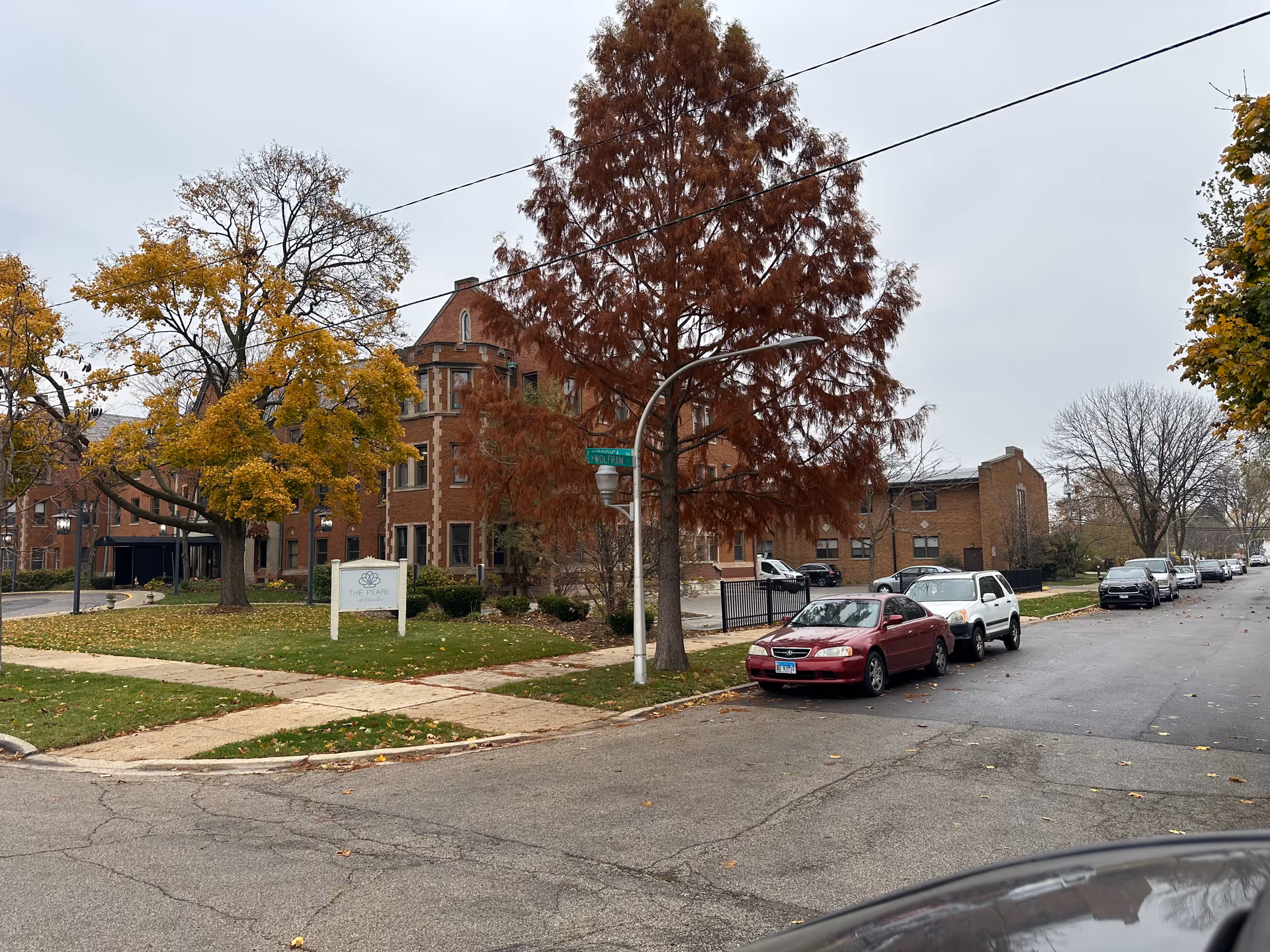 Exterior view of a brick building with a sign in front that reads 'The Pearl of Montclare'. The scene includes a street corner with parked cars, trees with autumn foliage, and a cloudy sky.