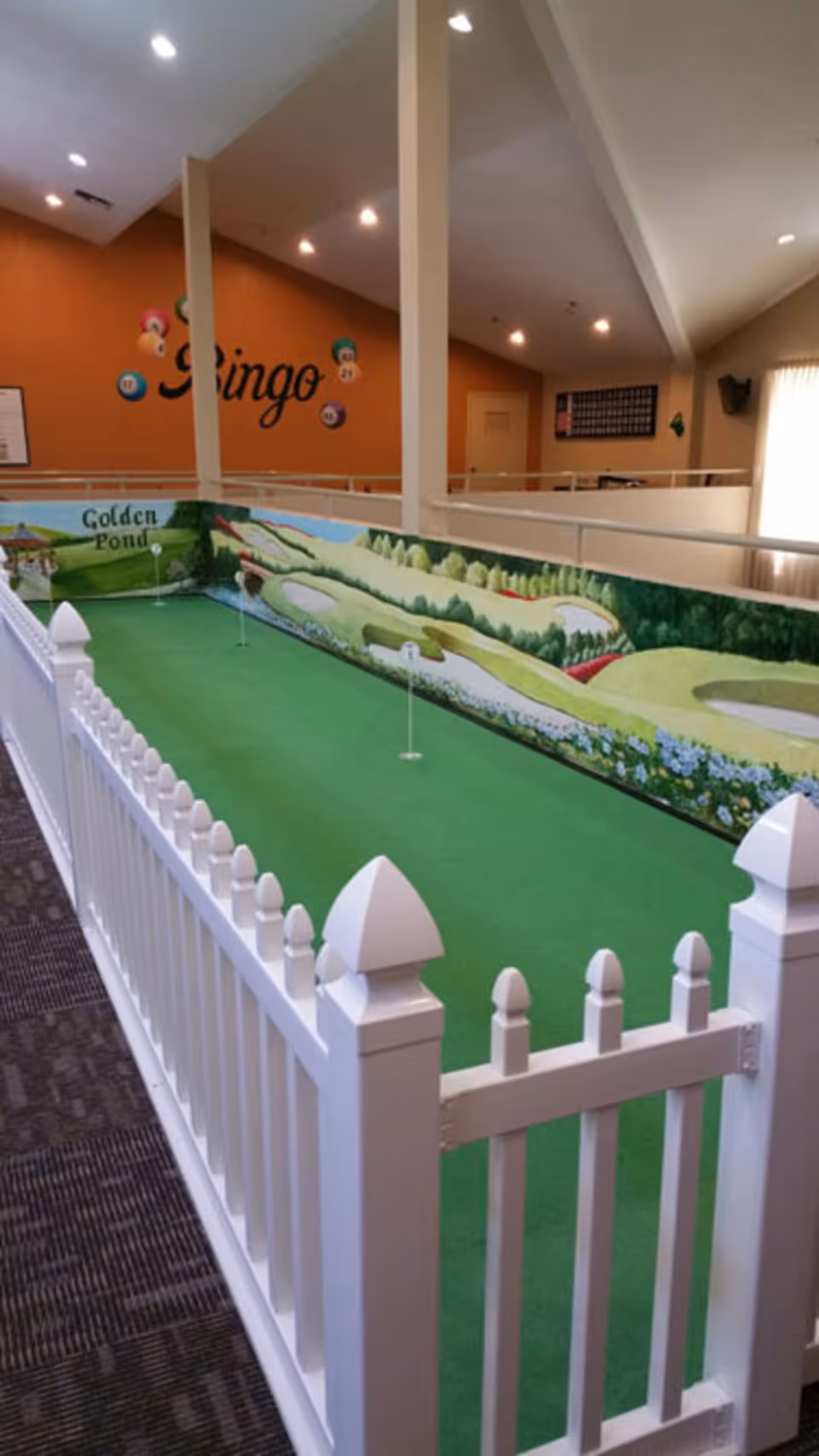 Indoor miniature putting green enclosed by a white picket fence with a mural of a golf course on the wall. The area is well-lit with ceiling lights and has an orange wall with the word 'Bingo' and bingo balls displayed.