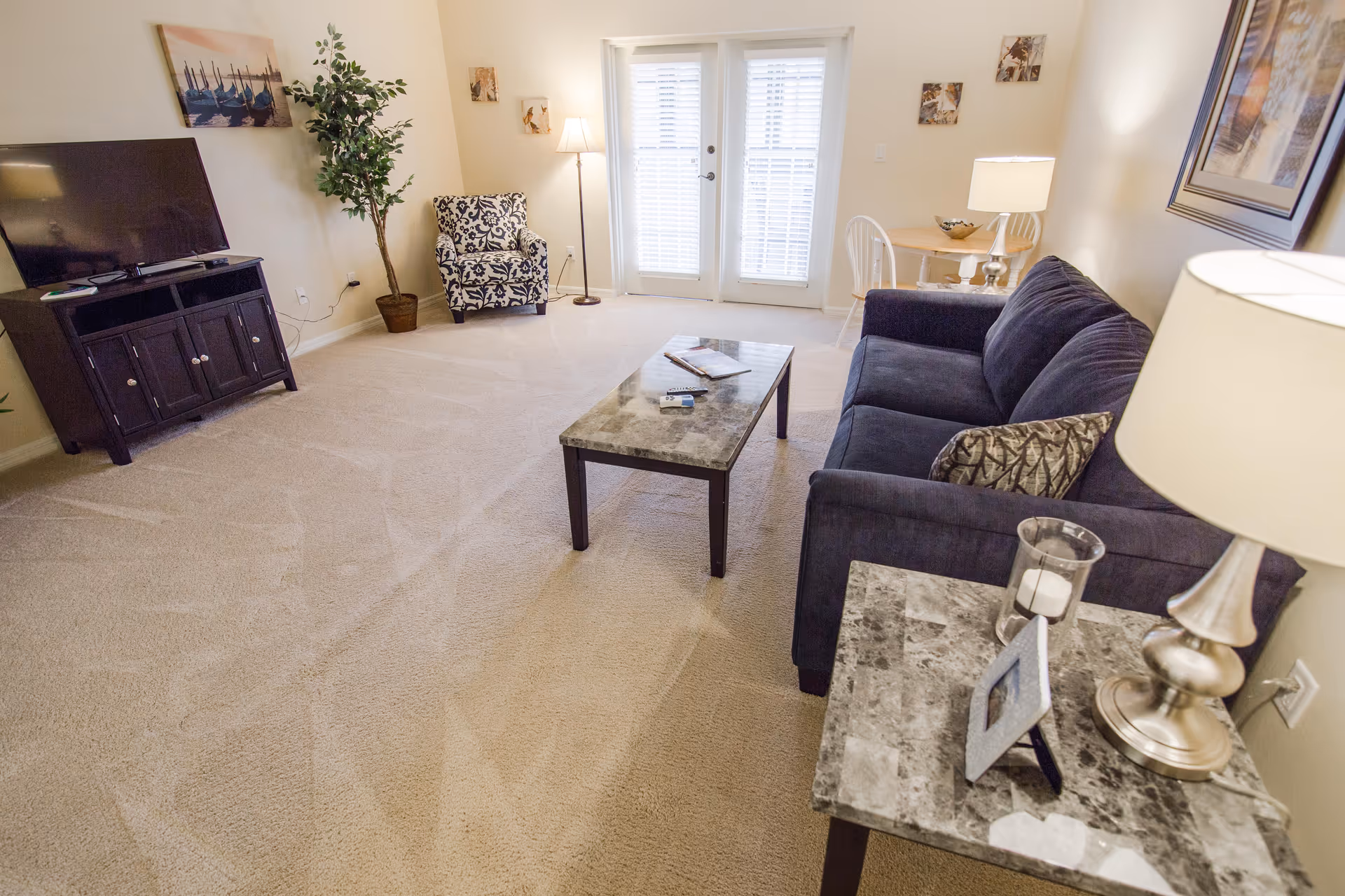 Bright furnished living room with a dark sofa, marble-topped coffee and side tables, TV console, armchair, and French doors.