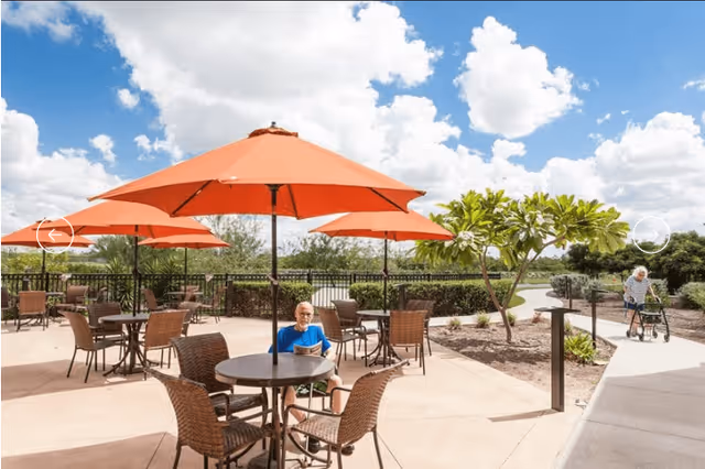 Outdoor patio area with several round tables and orange umbrellas. A man is sitting at one of the tables reading a newspaper, and an elderly woman with a walker is walking on a nearby pathway. The area is surrounded by greenery and a partly cloudy sky is visible.