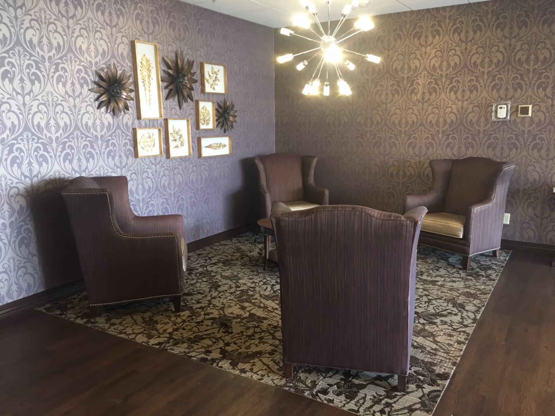A cozy sitting area with four brown upholstered armchairs arranged around a small round wooden table on a patterned rug. The walls have a purple damask wallpaper with framed botanical art and decorative metal wall sculptures. A modern chandelier with multiple light bulbs hangs from the ceiling, illuminating the room.