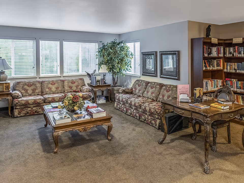 A cozy living room area in a senior living facility with two floral-patterned sofas, a wooden coffee table with books and a flower arrangement, a wooden side table with a lamp, a large potted plant in the corner, framed pictures on the wall, and a wooden desk with more books and a chair. Bookshelves filled with books are visible in the background.